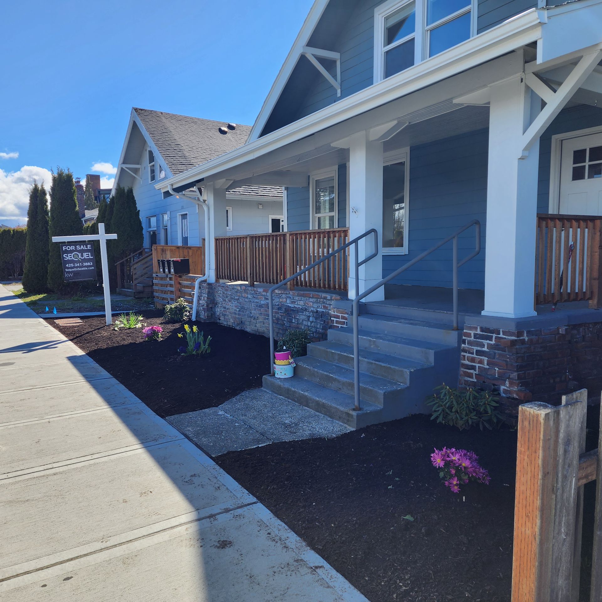 Blue house with porch and steps, surrounded by fresh mulch and flowers on a sunny day.
