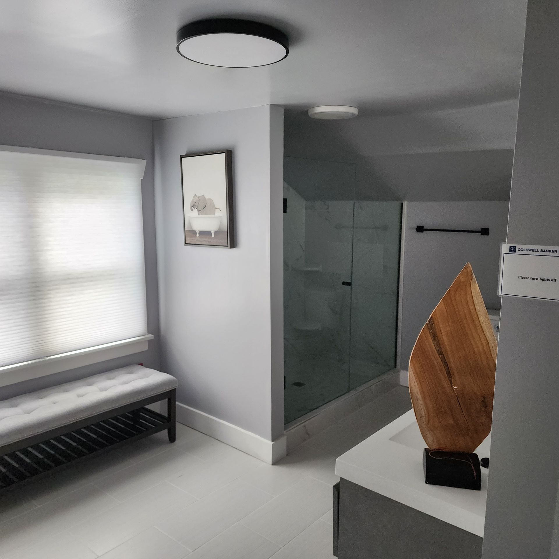 Bathroom with bench, framed art, shower, and decorative wooden sculpture. Gray walls, white trim and tile.