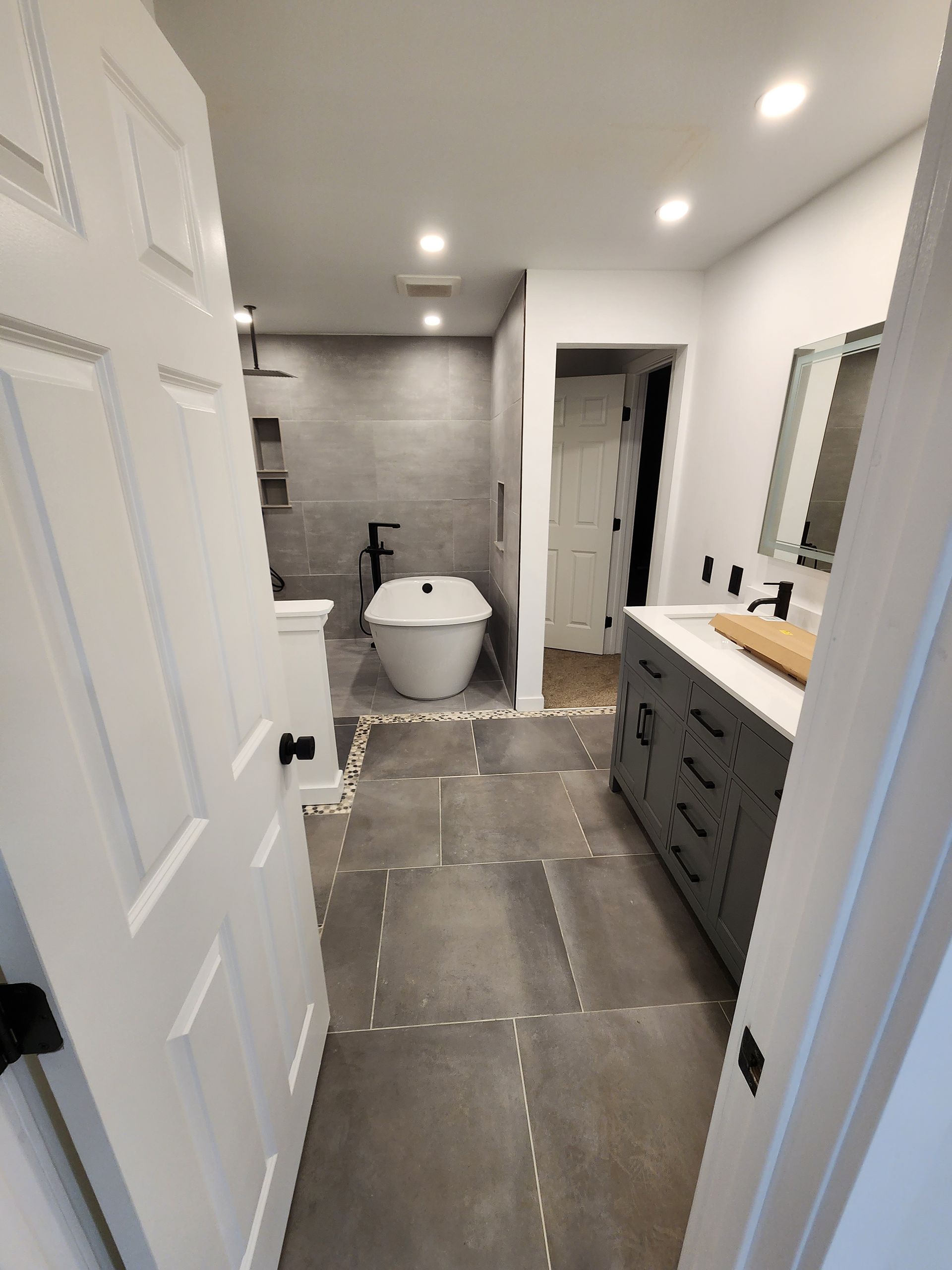 Bathroom with gray tile, freestanding tub, gray vanity, white doors, and black fixtures.