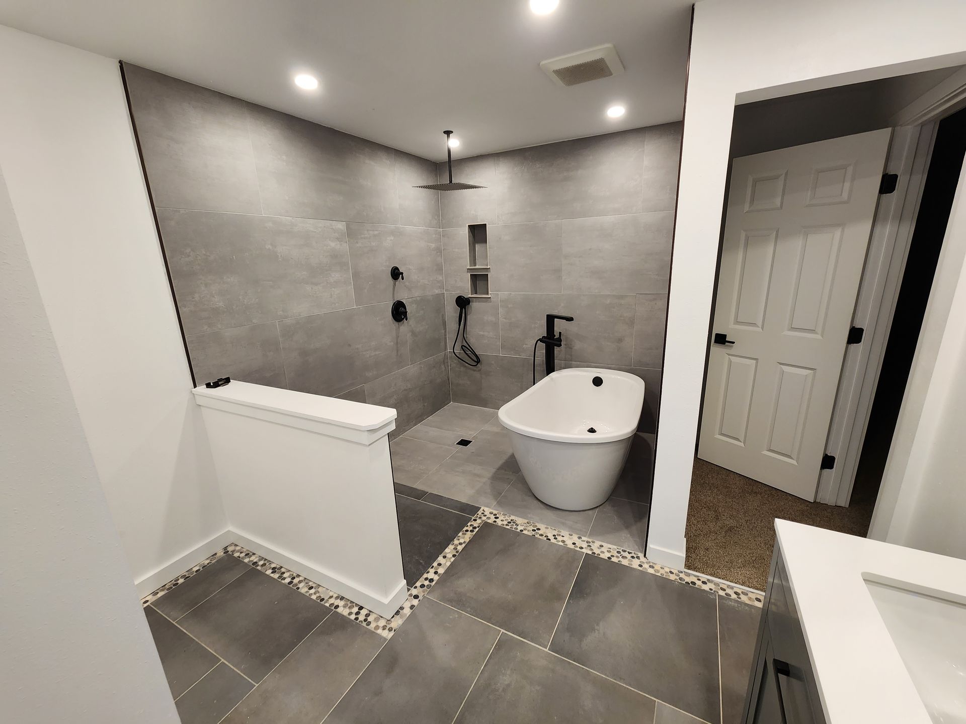 Modern bathroom with gray tile, walk-in shower, freestanding tub, and white door.