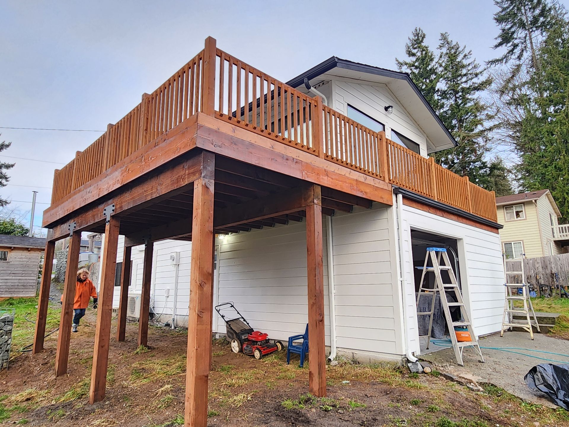 Wooden deck on a two-story building. A person walks below. Lawn mower sits nearby. Overcast sky.