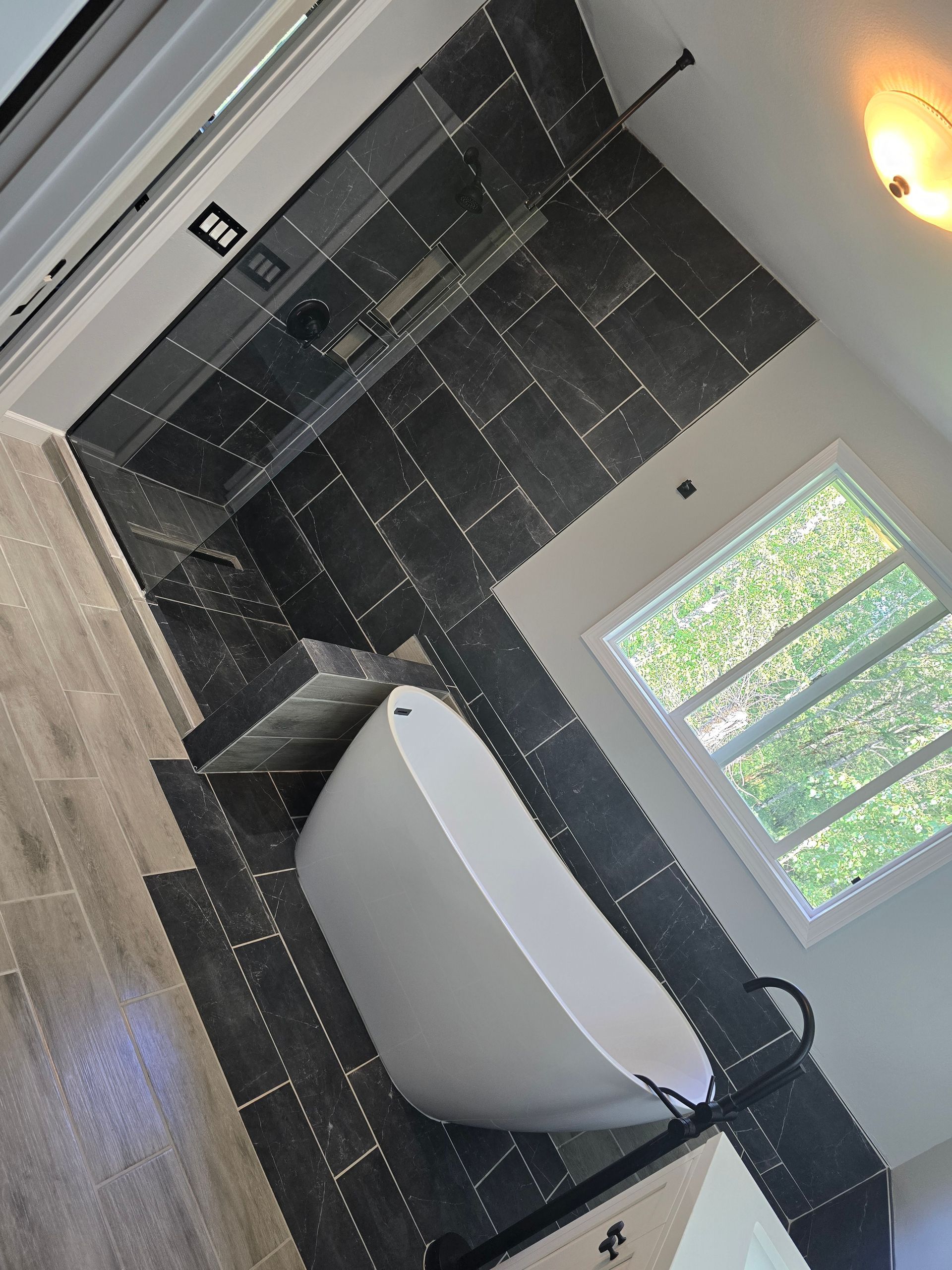 Modern bathroom with gray tiled shower and floor, a white tub, and a window.