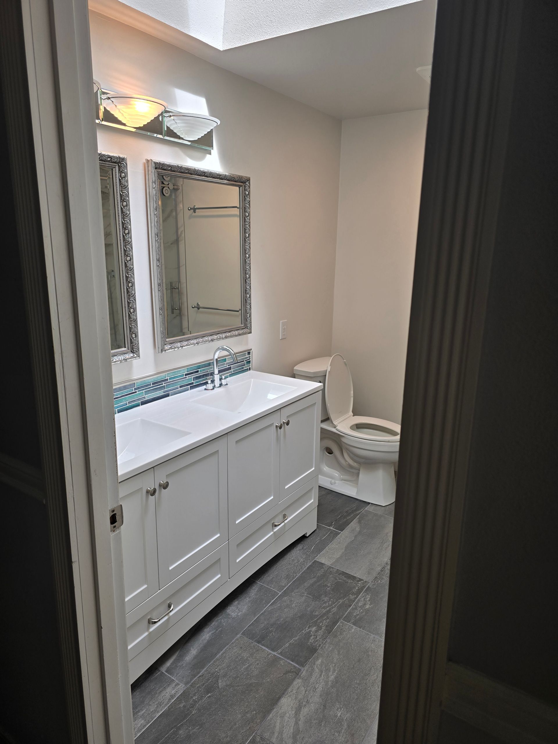 Bathroom with white vanity, gray tile floor, toilet, and mirrors. Natural light from a skylight.