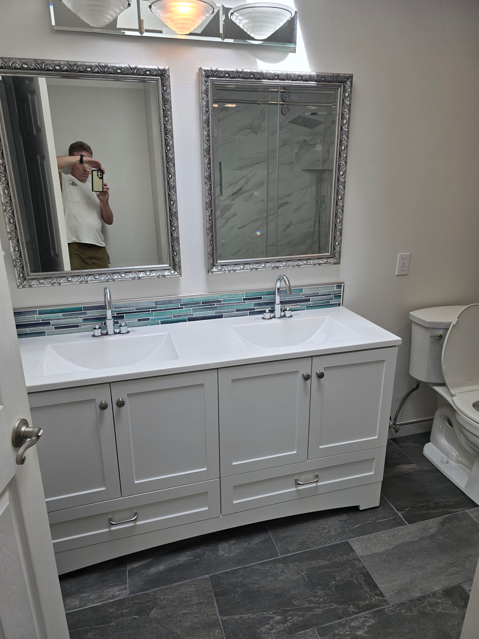 Bathroom with white double vanity, mirrors, blue tile, toilet, and gray floor tiles.