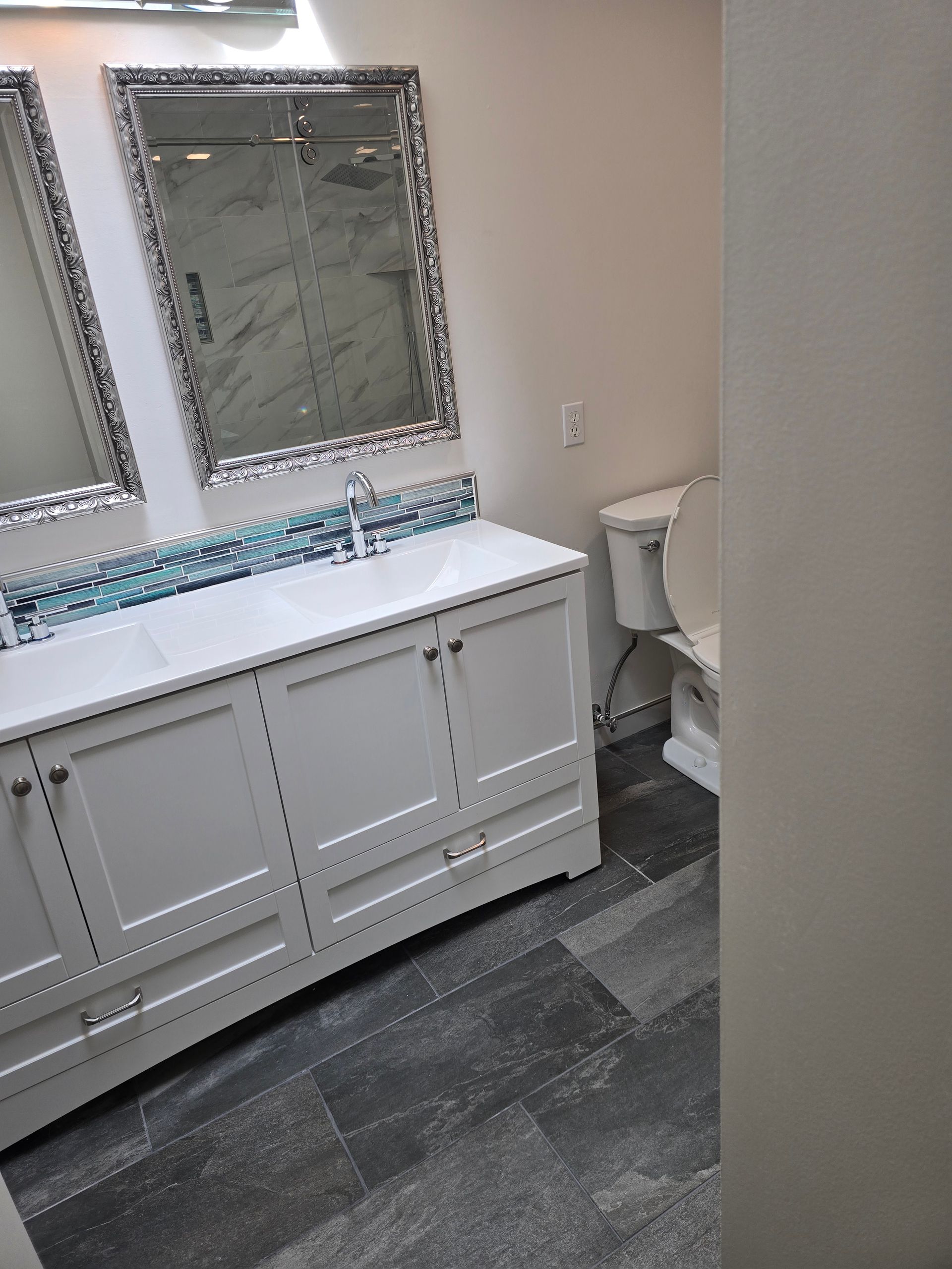 Bathroom with white vanity, blue tile backsplash, two mirrors, and a toilet. Gray tile floor.