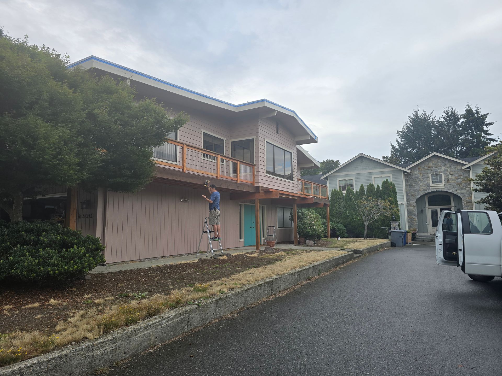 Person painting a two-story beige house with a wooden deck. Cloudy sky. A van is parked nearby.