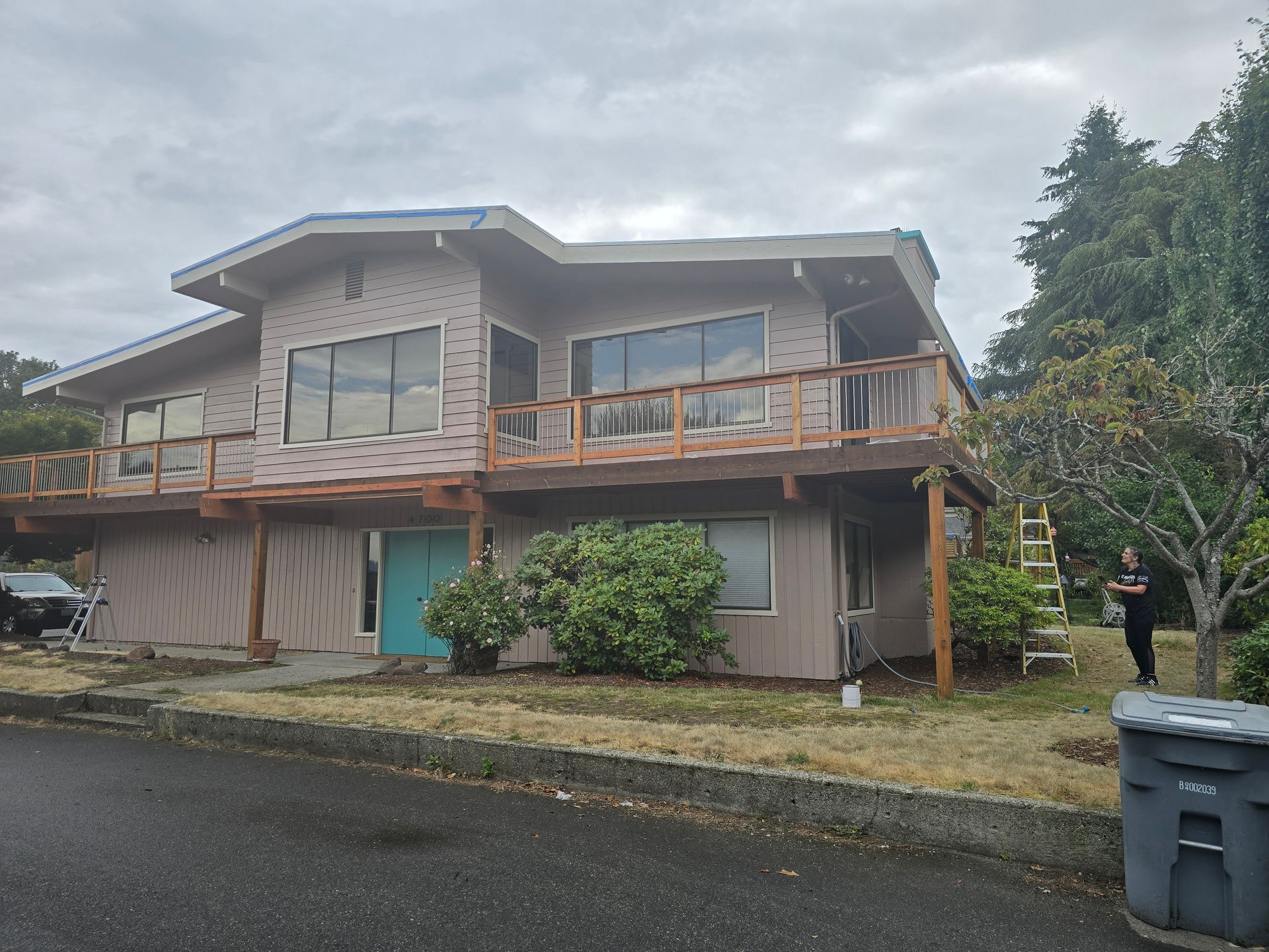 Two-story house with brown siding, decks, and a turquoise front door, person standing beside a ladder.