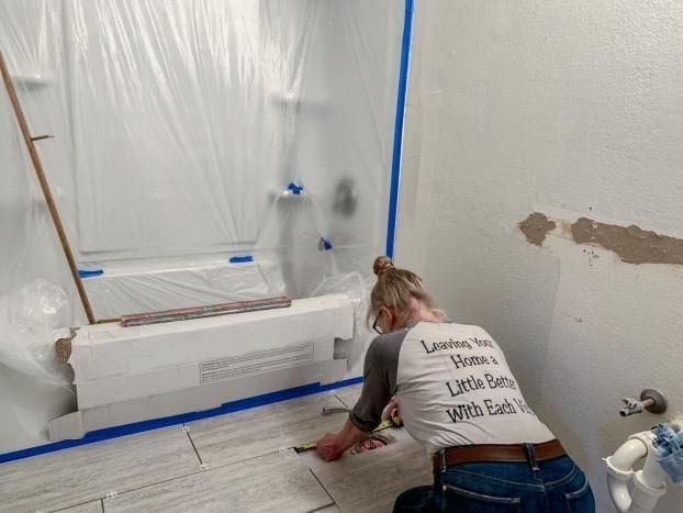 Woman tiling a bathroom floor. Walls are white with some damage. Tub and shower area is covered in plastic.