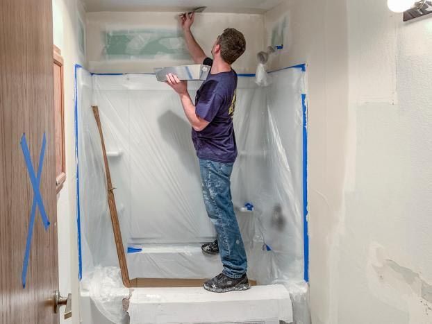 Person paints shower area, standing on a platform, masked, plastic covers everything, walls are taped.