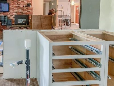 Kitchen renovation in progress: white cabinets with open drawers, plumbing, and a view of the adjacent room.
