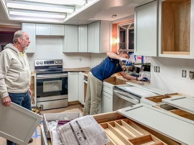 Two people installing kitchen cabinets. One holds a door; the other works near the sink, surrounded by cabinet parts.