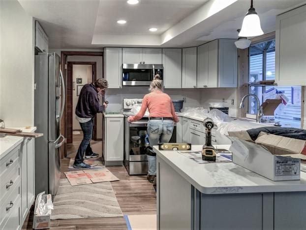 Two people installing new kitchen cabinets; light blue cabinets, grey countertops, appliances, construction tools.