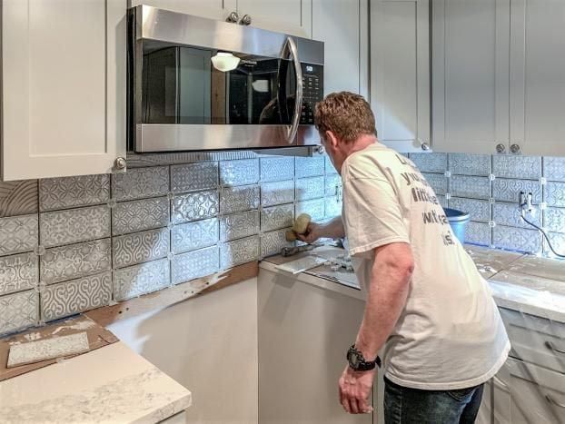 A person tiling a kitchen backsplash, applying grout with a sponge near a microwave.