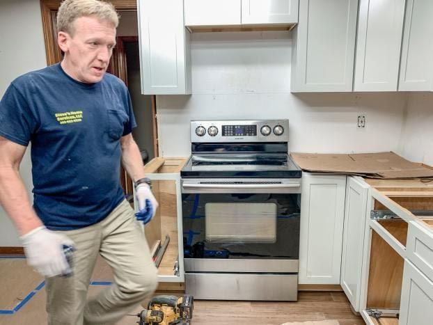 Man in a kitchen during renovation, wearing gloves, looking concerned. Cabinets, stove, and tools are visible.