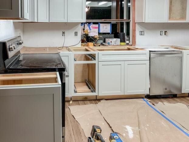 Kitchen renovation in progress: white cabinets, stove, dishwasher, wood countertops, and construction tools.