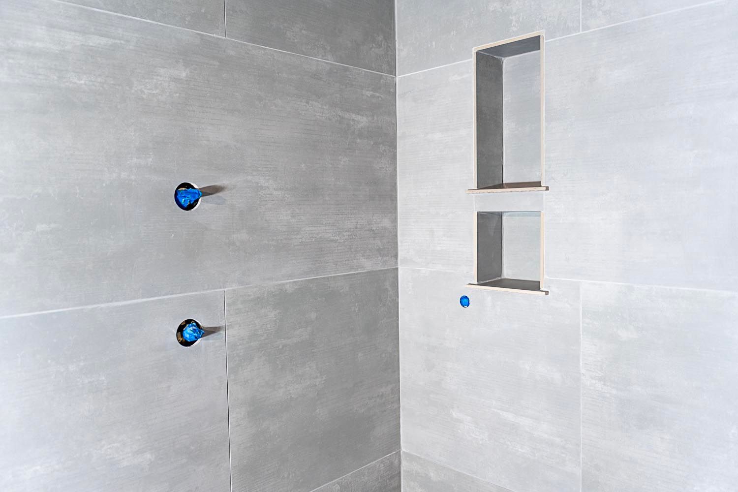 Gray tiled shower wall with blue pipe openings and a built-in shelf.