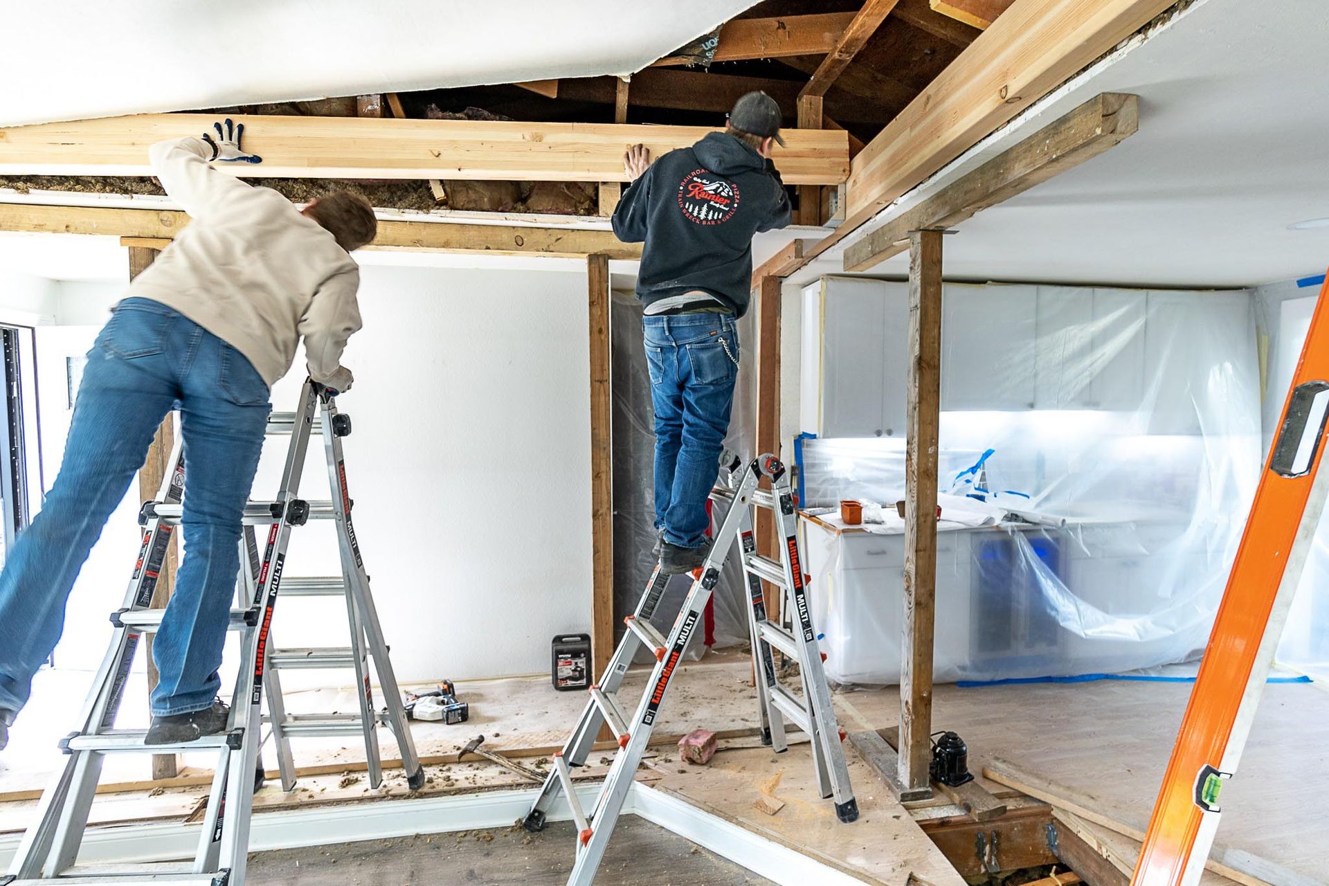Two people on step ladders work on a home renovation, installing wooden beams in a room.