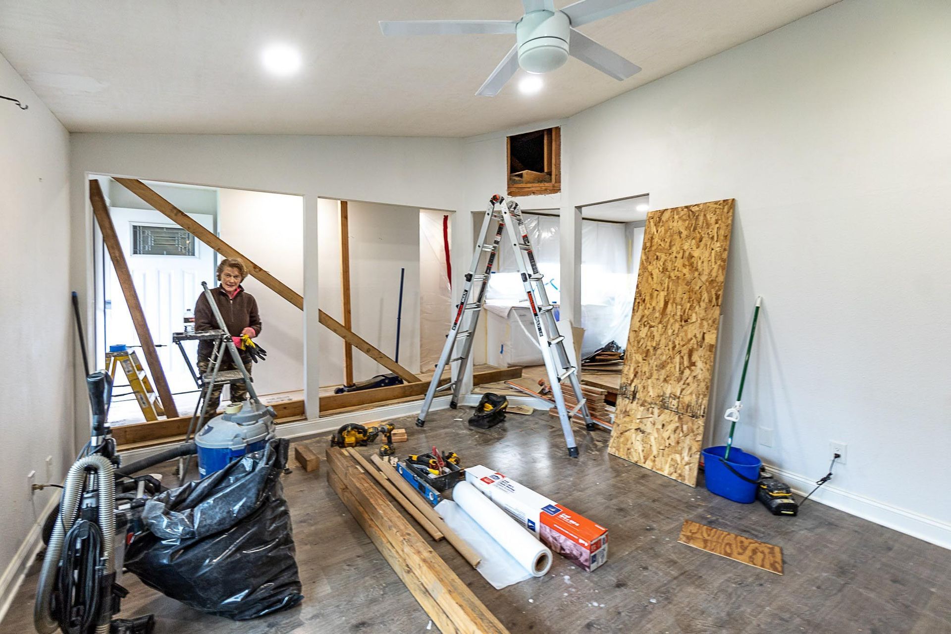 A person renovating a room, walls partially removed. Tools and materials are scattered on the floor.