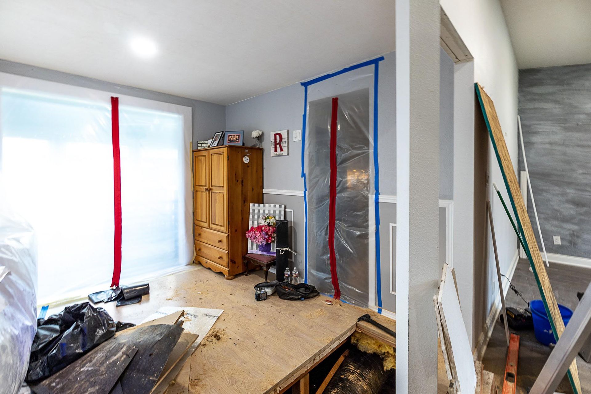 Room under construction. Plastic sheeting taped to doorways, debris on floor. Wooden dresser and chair visible.