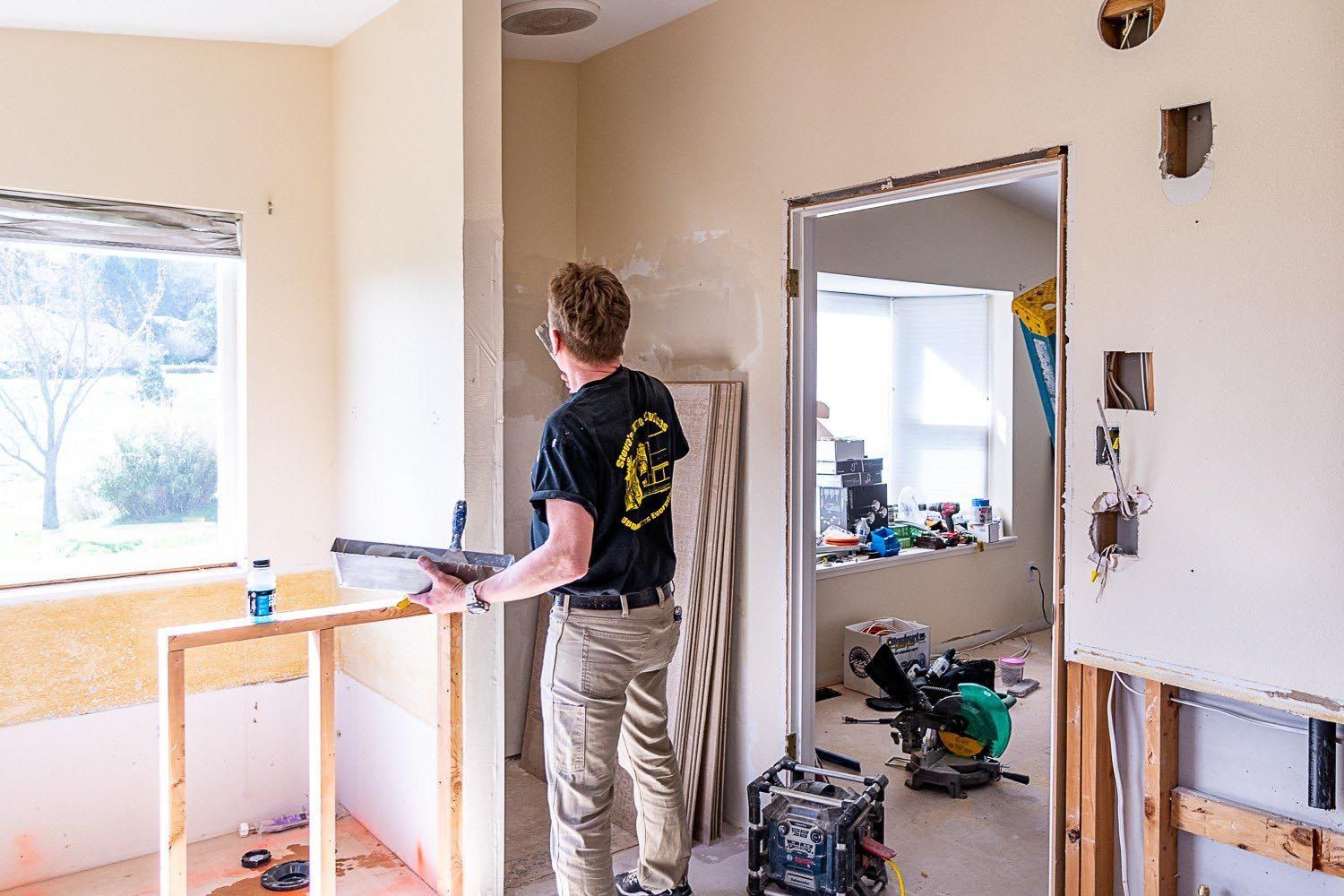 Man renovating a room, holding a tool, surrounded by construction materials.
