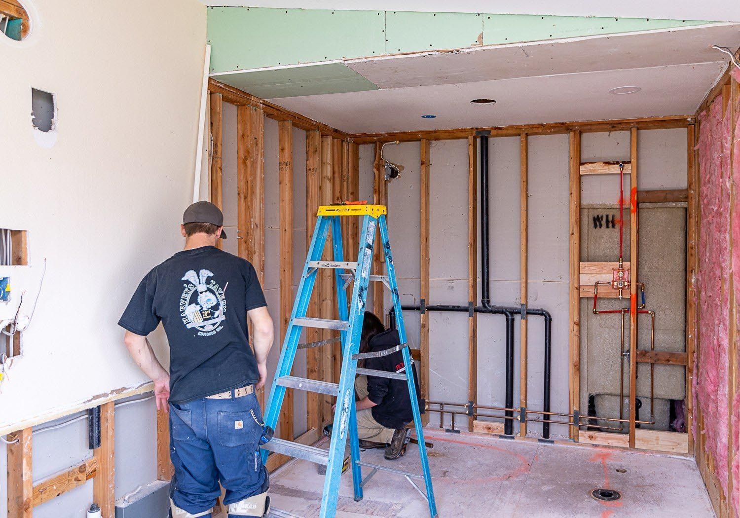 Two people working on a room's renovation. One on ladder, other standing. Exposed studs, pipes, insulation visible.