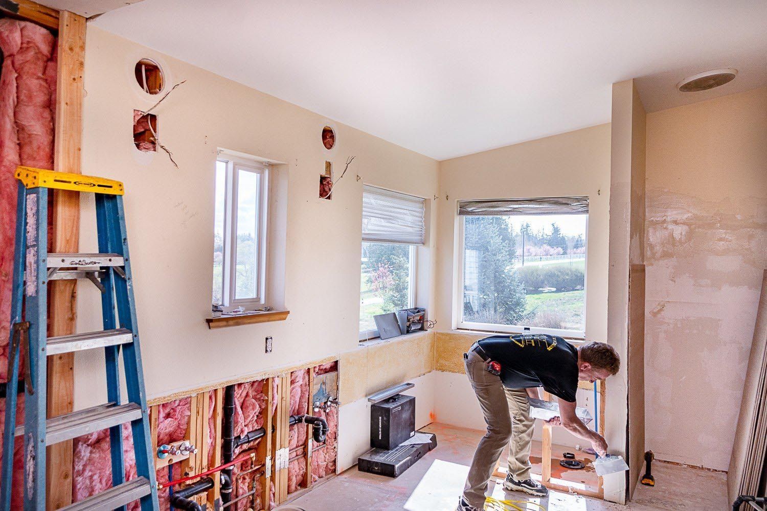 Person renovating a room; exposed wall studs, person painting, ladder, windows, pink insulation.