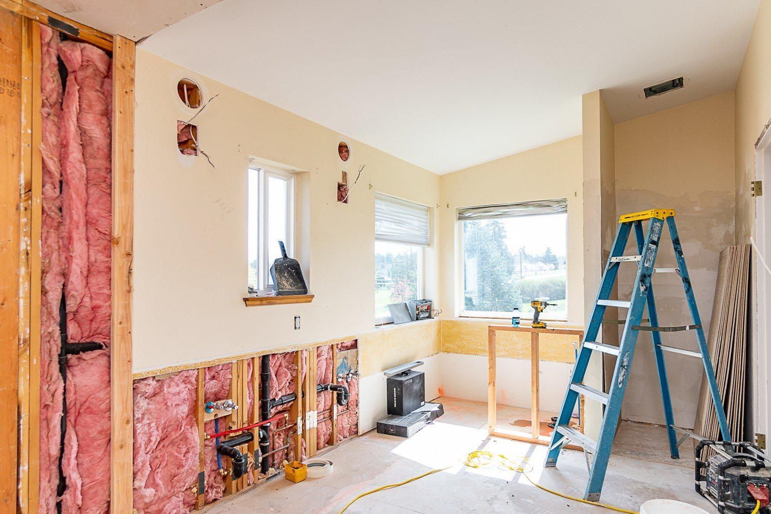 Interior room under renovation, exposed framing, insulation, electrical work. Ladder, windows, beige walls.