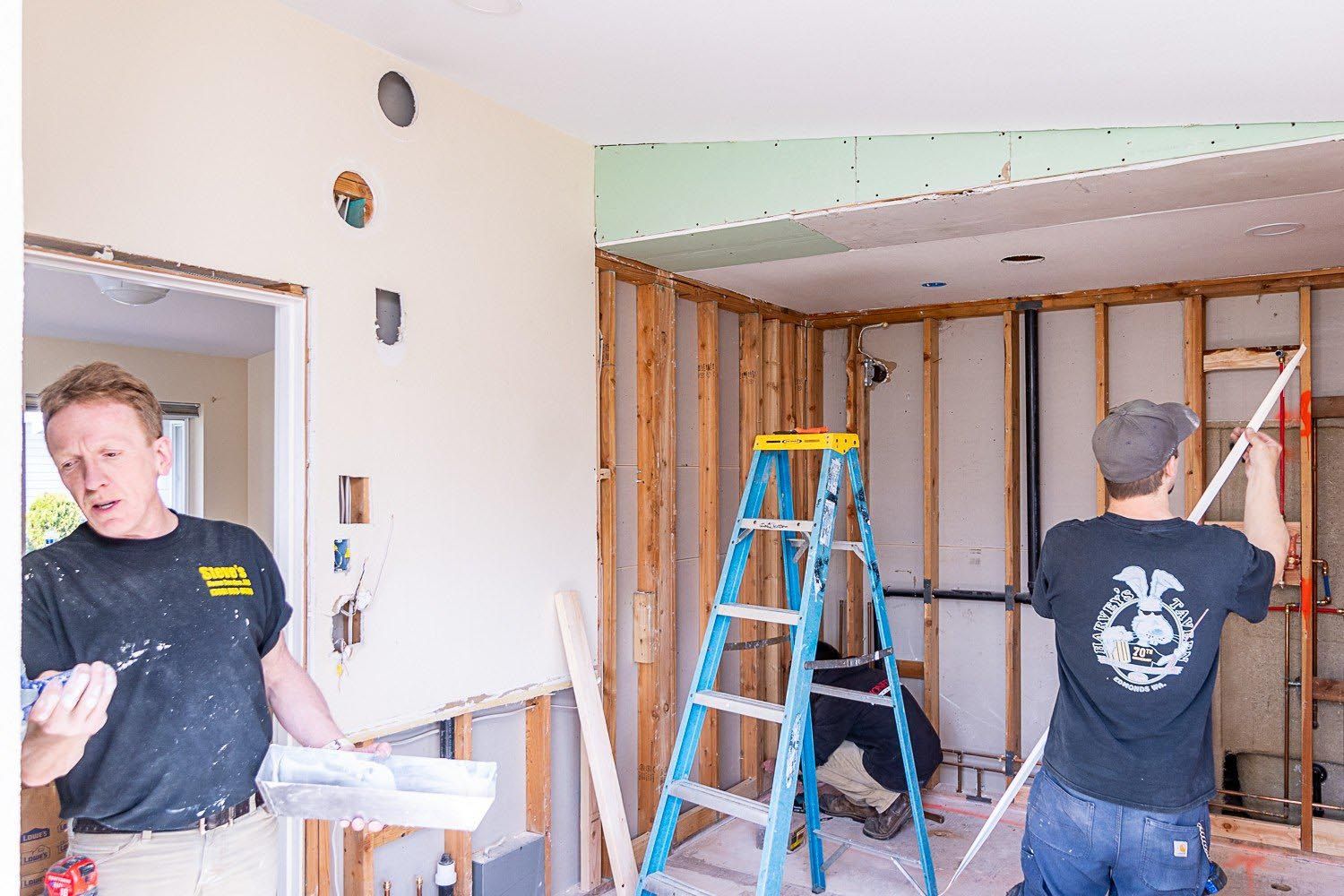 Two men renovating a room, one holding tools, the other measuring. Interior shows exposed framing, a ladder, and drywall.