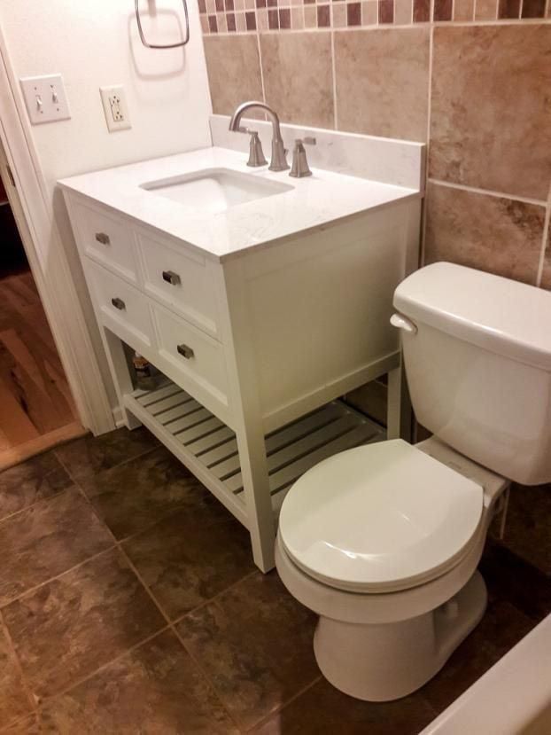 White vanity with sink, two drawers, and open shelf next to a white toilet in a bathroom with tile.