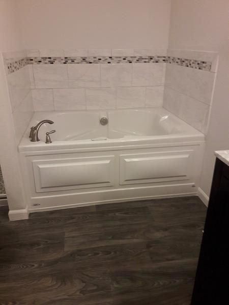 White-tiled bathtub with mosaic accent, white cabinetry, chrome faucet, and dark wood-look flooring.