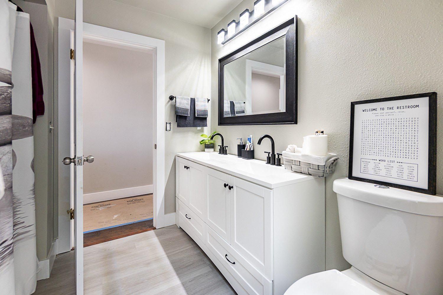 Bathroom with white vanity, black framed mirror, and toilet, with a towel rack and open doorway visible.