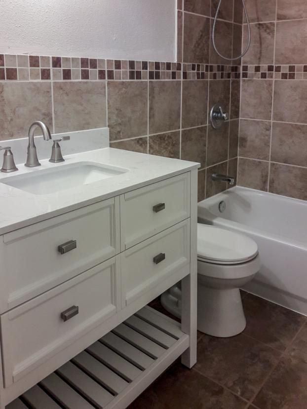 White bathroom vanity and toilet, brown tiled walls, and a bathtub.