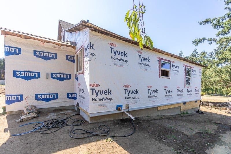 House under construction, covered in Tyvek wrap, windows visible. A tree is being lifted above.