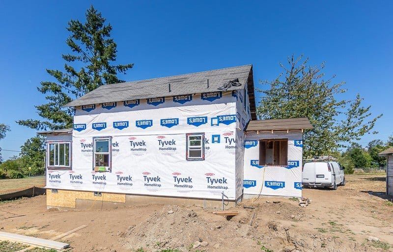 House under construction, wrapped in blue and white Tyvek, windows installed, brown roof, sunny day.