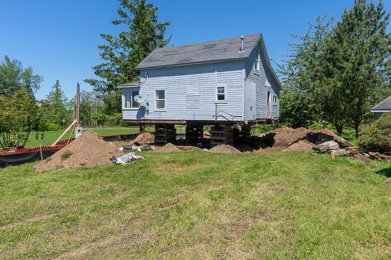 House raised on supports, likely for foundation work, on a grassy plot with piles of dirt under a blue sky.