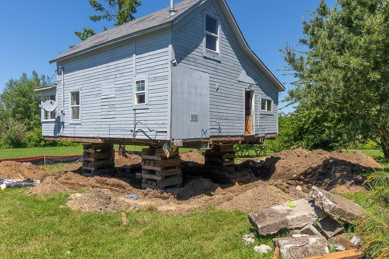 House lifted on wooden supports during foundation work. Blue siding, green grass, clear sky.