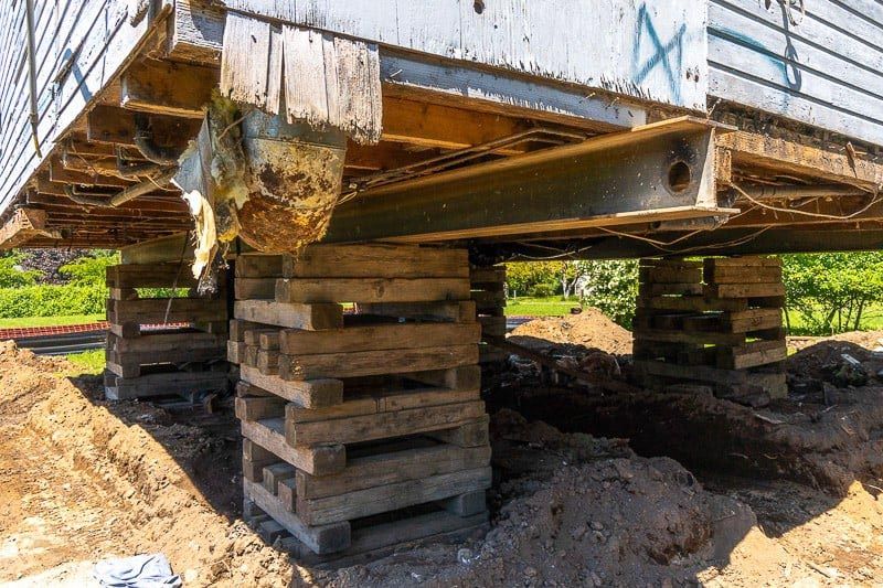 House raised on wooden supports during foundation work in dirt.
