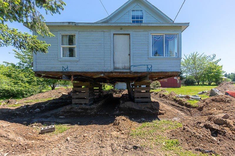Small, light-blue house raised on wooden blocks, likely for foundation work, in a grassy area.