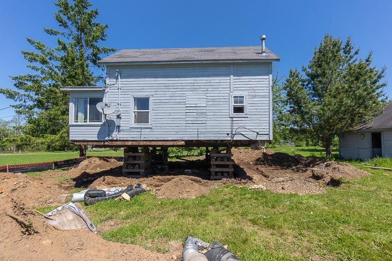 A small weathered house on wooden supports over a dirt excavation; sunny, outdoor setting.