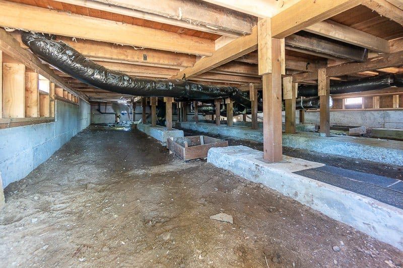 Underneath a raised wooden house, crawlspace with beams, ductwork, and concrete supports.