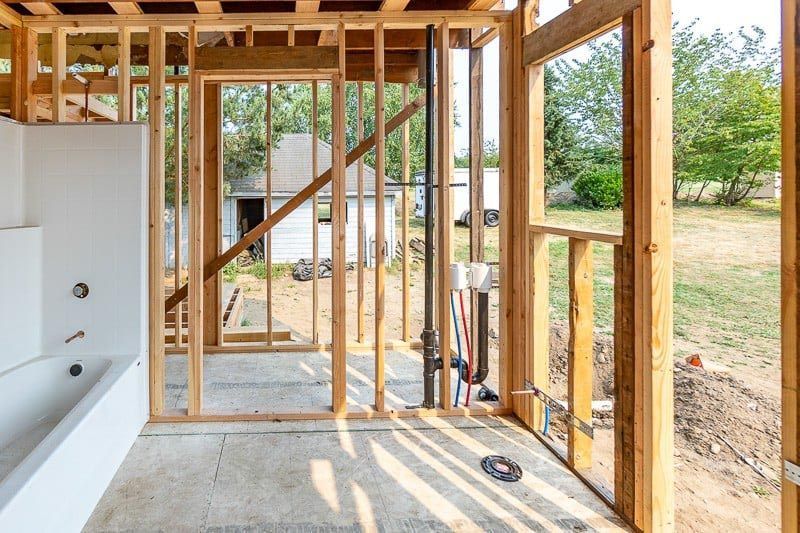 Interior view of a bathroom under construction with exposed wooden framing and a bathtub. Outside view through the window.