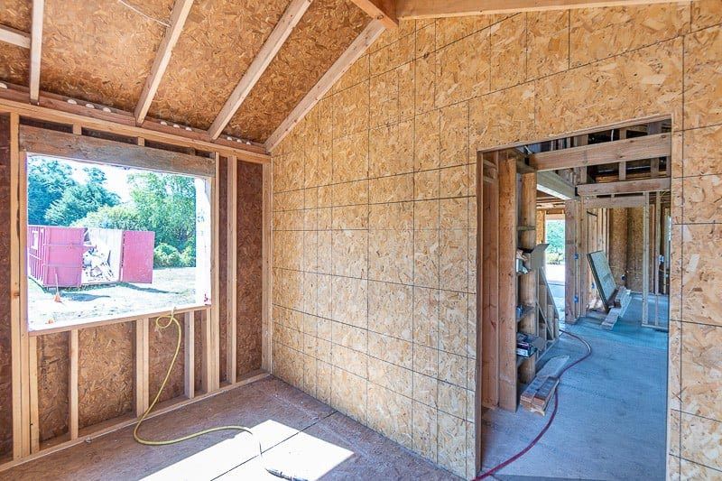 Interior of a room under construction, with exposed wood framing and OSB sheathing. Window and doorway visible.