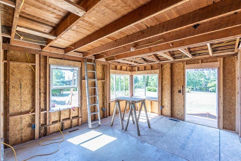 Interior of a room under construction, with wooden frame, windows, door, ladder, and sawhorses.