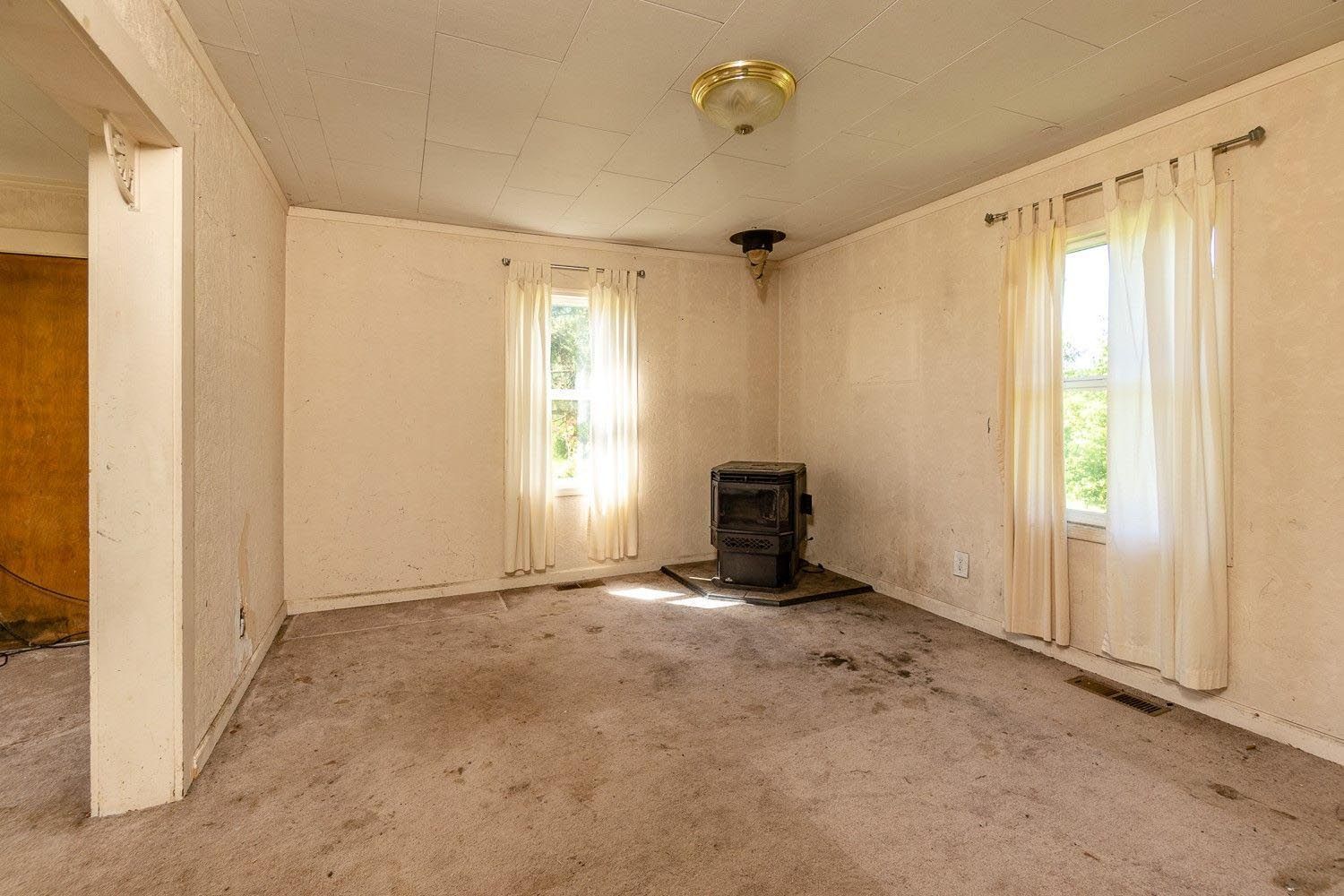 Empty room with faded walls, two windows with curtains, and a stove.