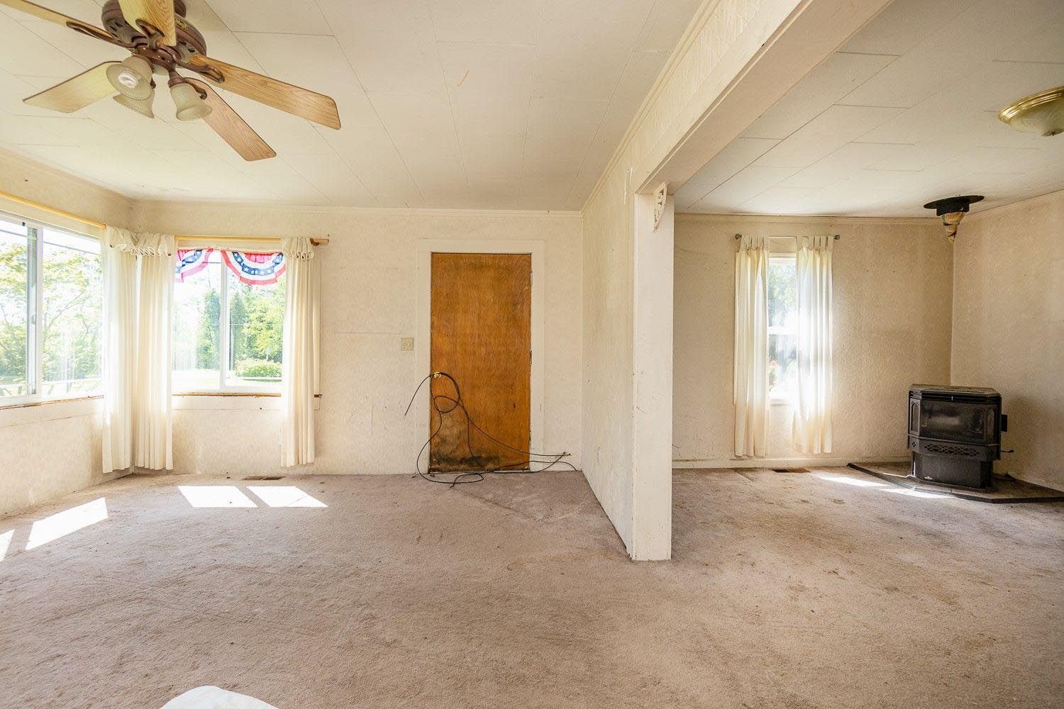 Empty, sunlit room with beige carpet, a ceiling fan, windows with sheer curtains, and a wood door.