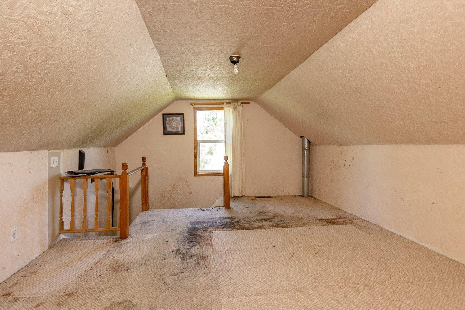 Attic room with sloped ceiling, window, carpet, and wooden railing, showing signs of wear and tear.
