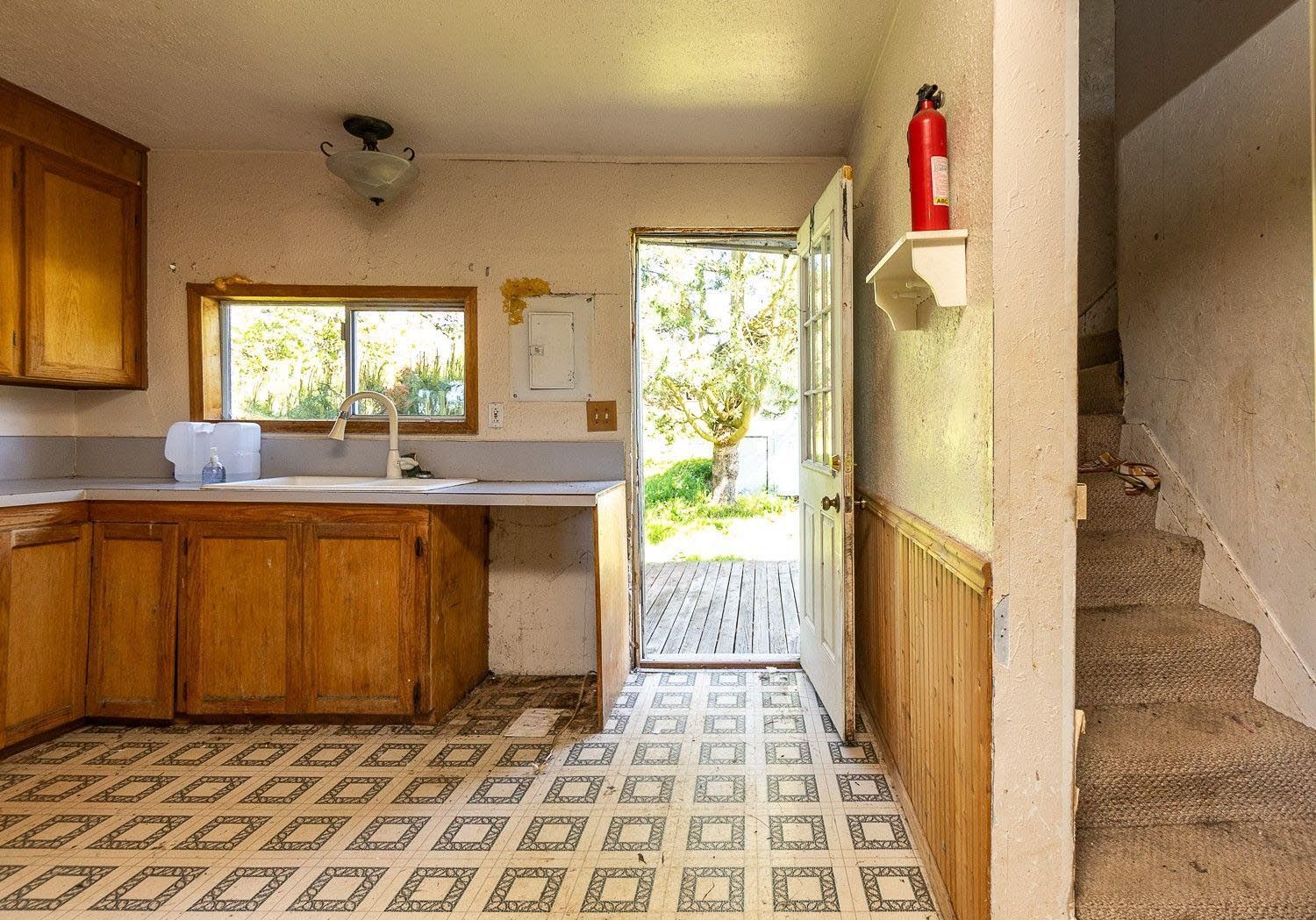 Small kitchen with wood cabinets, patterned floor, open door to outside, and stairs on the right.