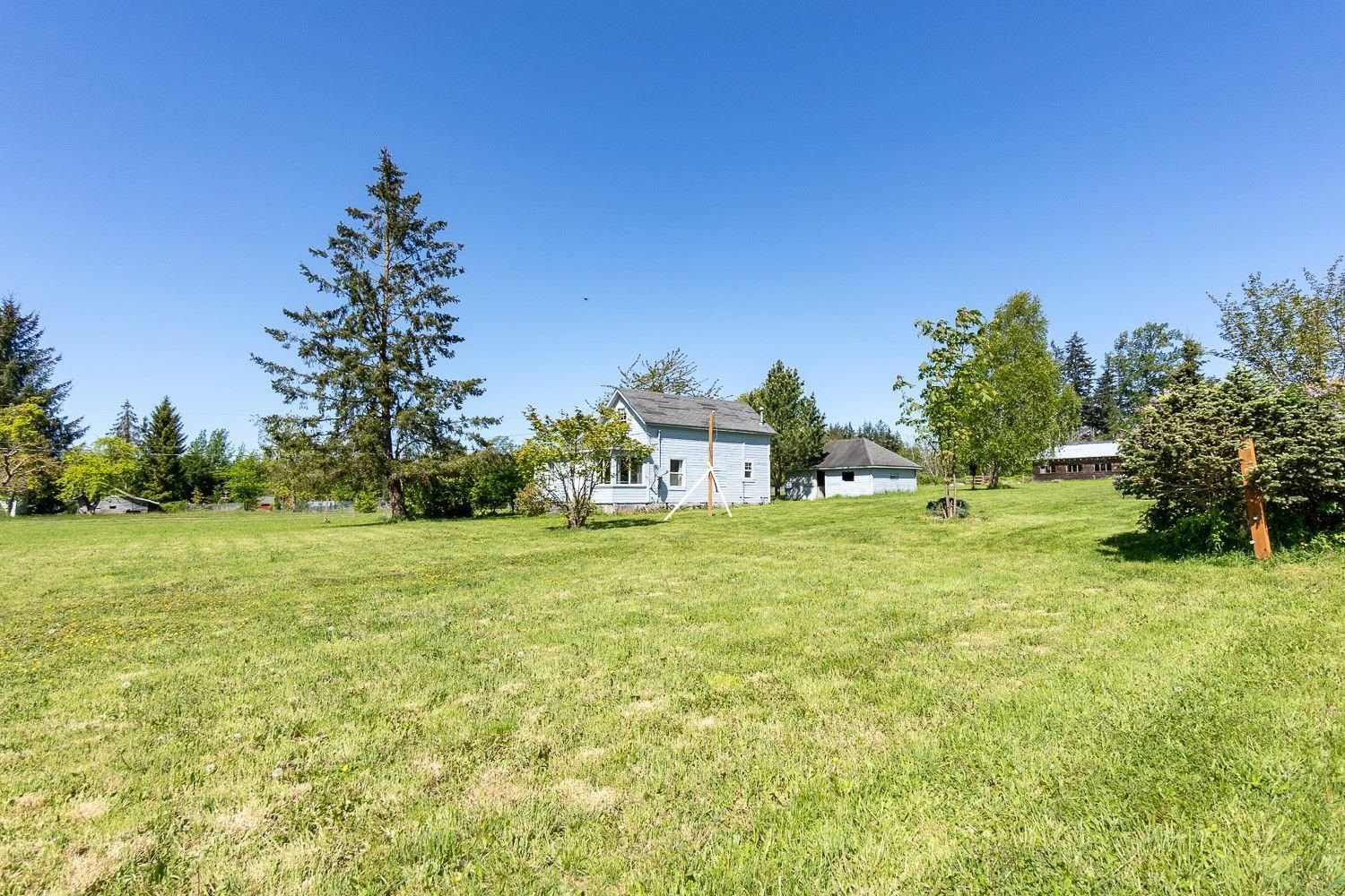 Grassy field with a light blue house and other buildings, under a clear blue sky.
