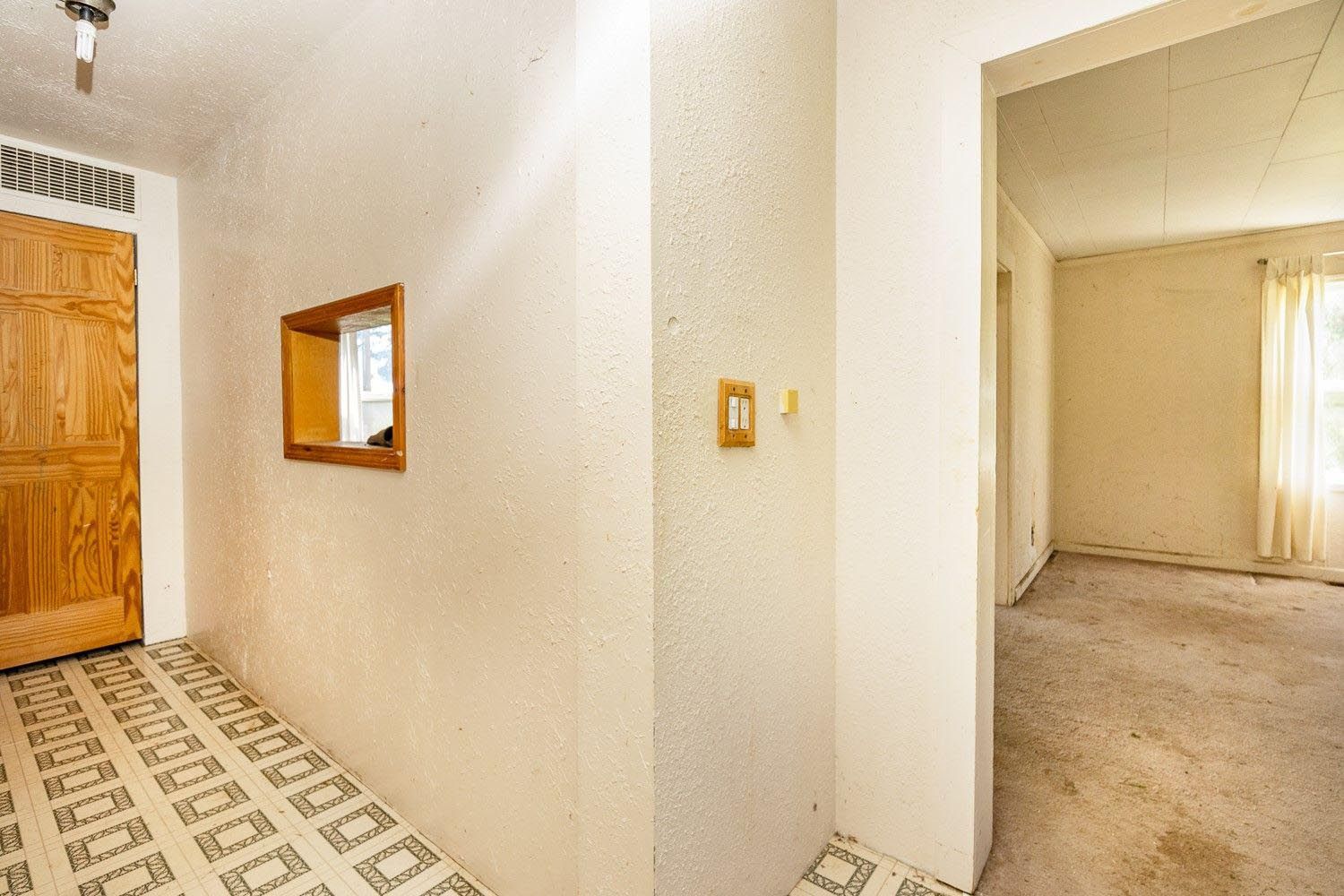 Hallway with tan door, square window, and view into room with window and beige curtains.