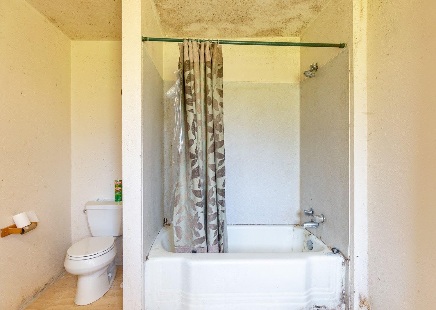 A small bathroom with a toilet, shower/tub, and a camouflage shower curtain. The ceiling has water damage.